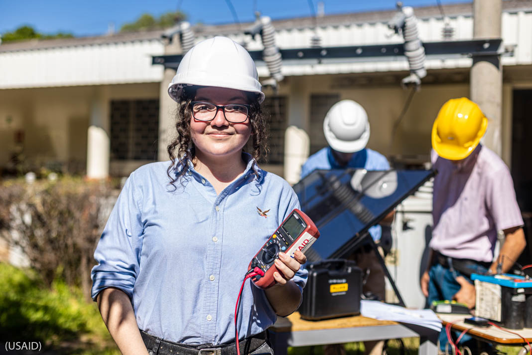 Woman in hard hat holding electrical instrument with "USAID" label, standing in front of two men in hard hats (USAID)