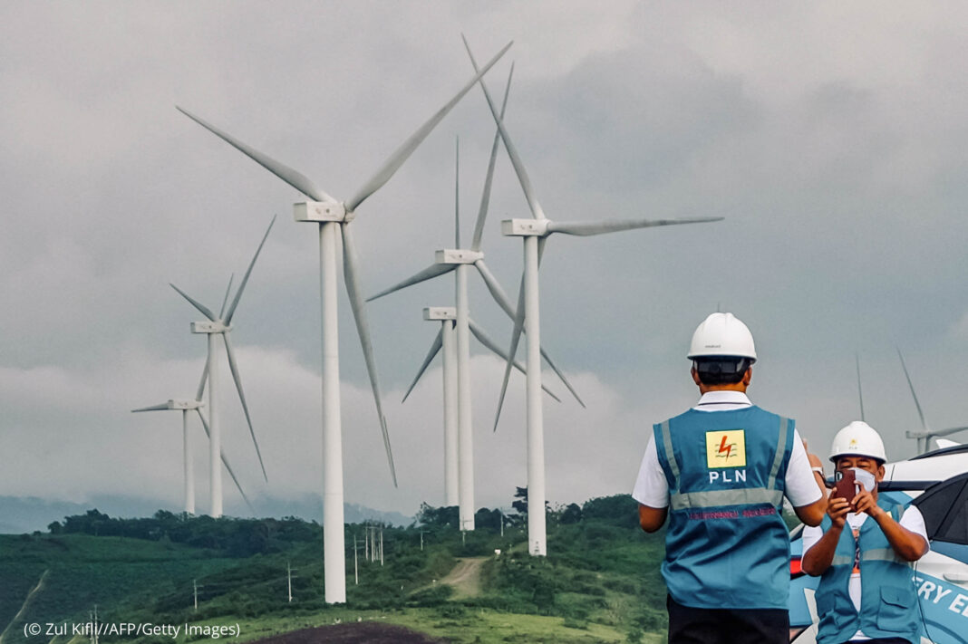 Man in hard hat and vest taking photo of similarly dressed colleague as they stand next to large wind turbines (© Zul Kifli//AFP/Getty Images)