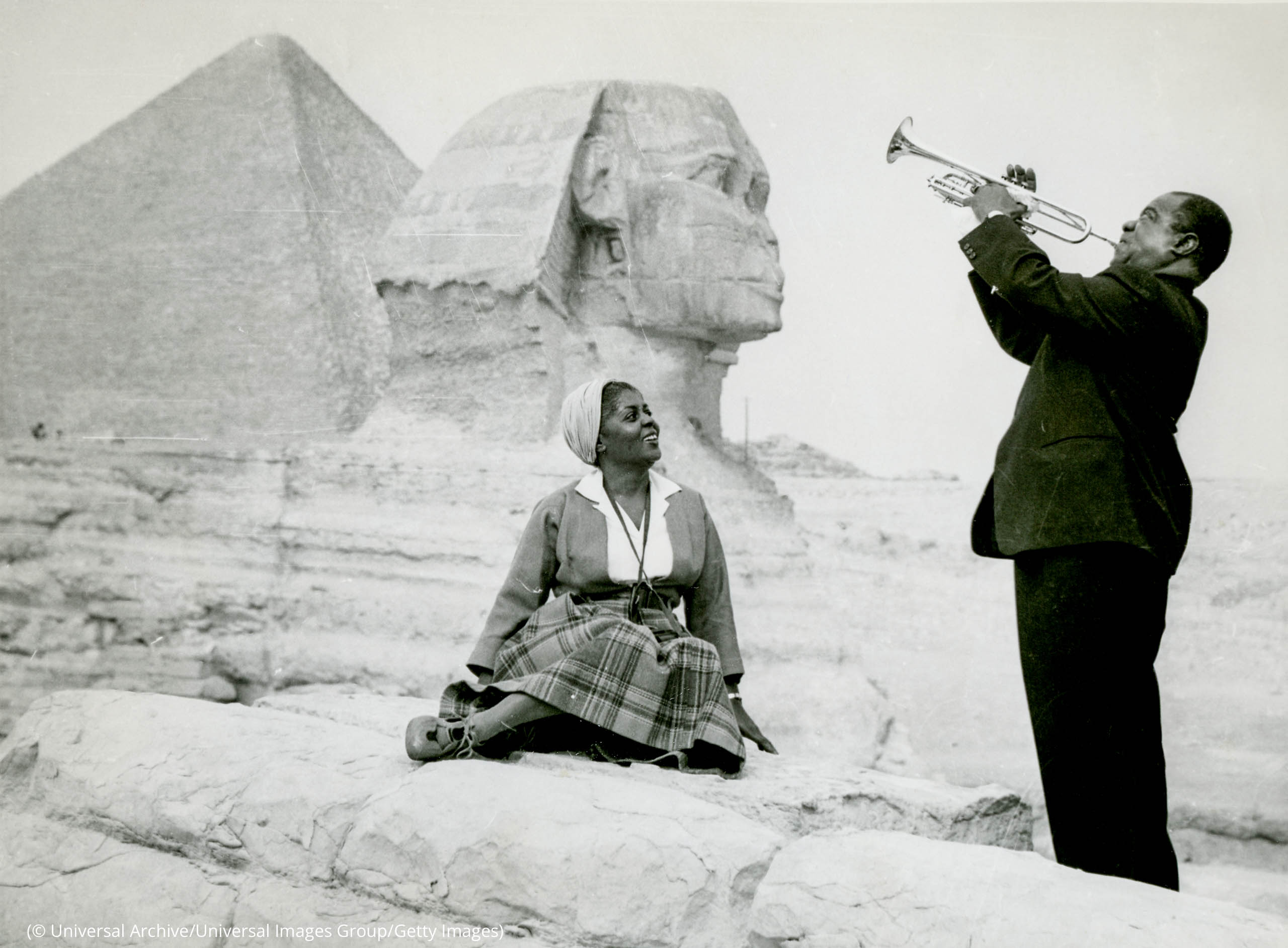 Louis Armstrong in suit playing trumpet in front of Sphinx with wife, Lucille, sitting on wall, watching (© Universal Archive/Universal Images Group/Getty Images)