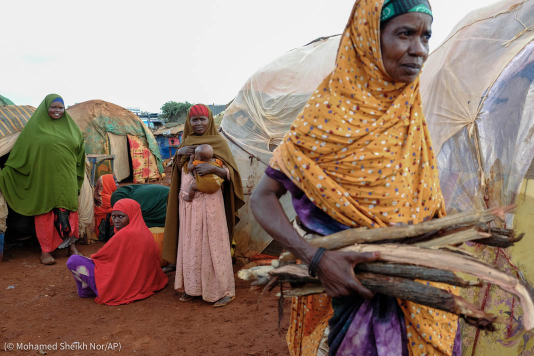 Woman carrying firewood past women and children standing and sitting next to tents (© Mohamed Sheikh Nor/AP)